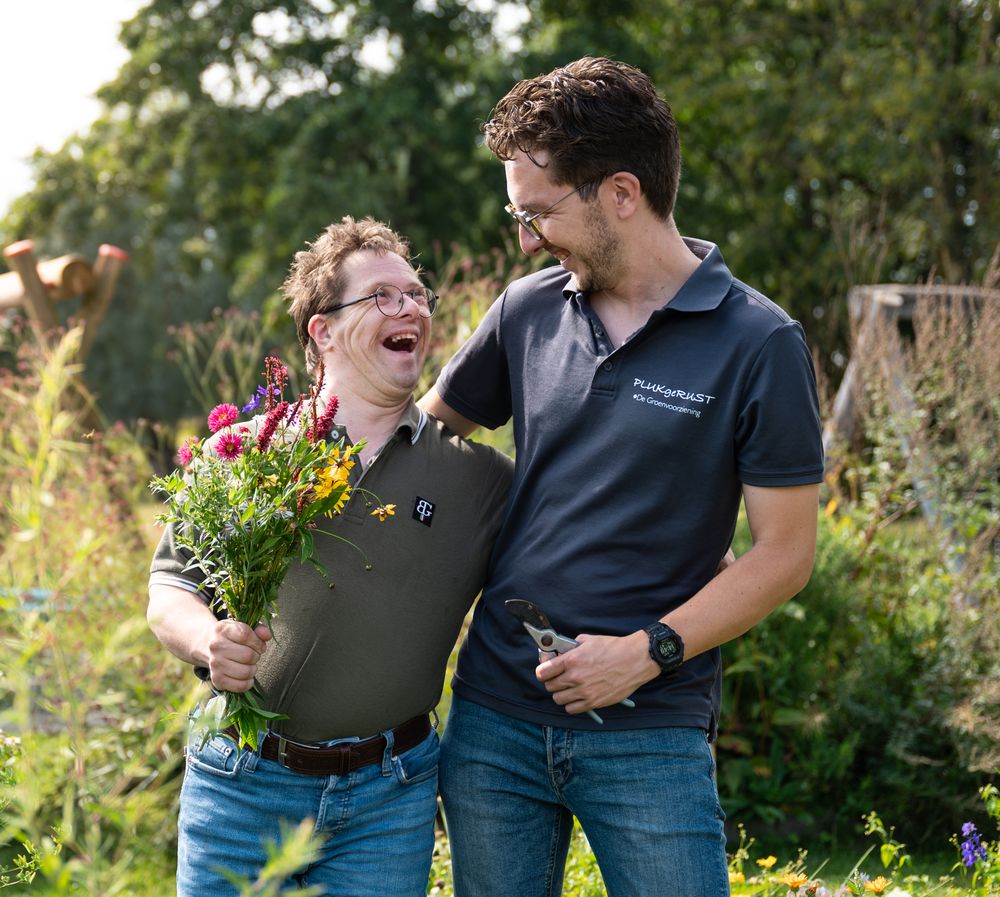 Een cliënt met een bos bloemen en een begeleider lachen samen in de tuin
